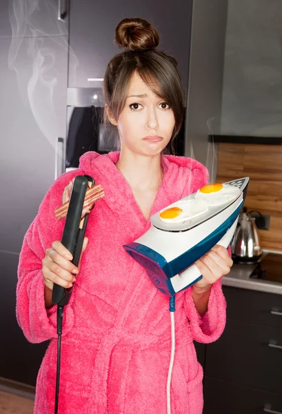 Beautiful young woman frying eggs and bacon on an iron and hair straightener — Stock Photo, Image