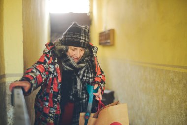 Üst düzey Doğu Avrupalı engelli kadın bir elinde mimari bariyerle binaya giriyor bir eliyle kapıyı açıyor, diğer eliyle koltuk değneğini tutuyor ayrıca sıcak ve pejmürde giyinmiş bir çanta tutuyor.