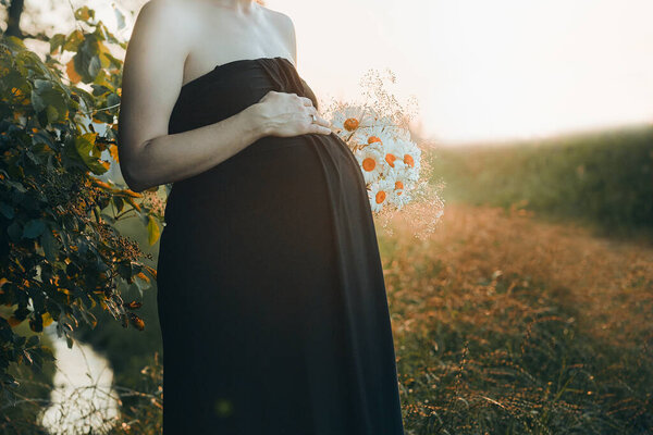 Pregnant woman hugging her tummy standing outdoors surrounded by nature. Pregnancy, expectation, motherhood concept