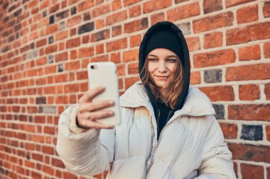Genç bir kadın video görüşmesi yapıyor, uzaktan konuşuyor, fotoğraf çekiyor, akıllı telefonu tuğla duvarın önünde duruyor.