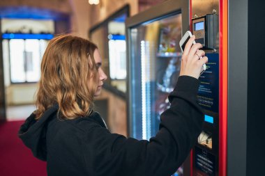 Otomatta ürün satın alan bir kadın cep telefonuyla ulaşılamaz ödeme metodu kullanıyor. Yeni ödeme yöntemleri kullanan bir kadın