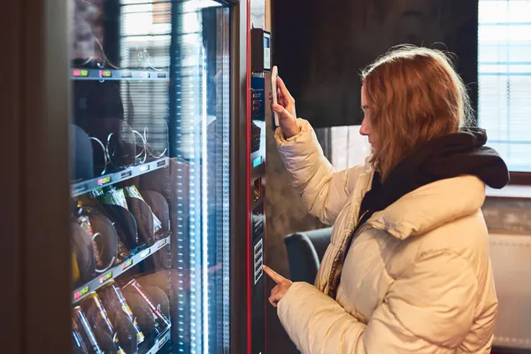 携帯電話で非接触で自動販売機で商品を支払う女性 新しい支払い方法を使う女性 — ストック写真
