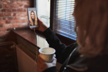 Genç bir kadın video görüşmesi yapıyor, uzaktan konuşuyor, akıllı telefon tutarak selfie çekiyor. Kahve içen kız ofiste dinlenirken pencerede duruyor.