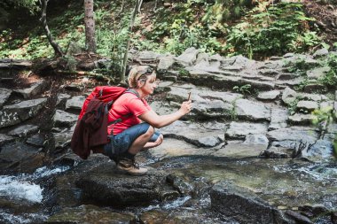 Tatilden fotoğraf çekiyordum. Sırt çantalı bir kadın yaz gezisi sırasında dağ akıntısı üzerinde duran akıllı telefon kamerasıyla manzaranın fotoğraflarını çekiyor.