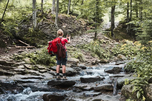 在度假的时候拍照夏天旅行时 背着背包站在山溪岩石上用智能手机拍照的女人 — 图库照片