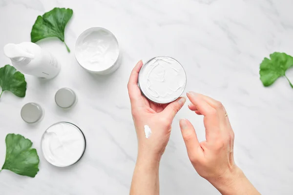Woman applying  cosmetic moisturizing hand cream. Cosmetic products, green leaves on white table. Spa, manicure, skin care concept. Flat lay, overhead view