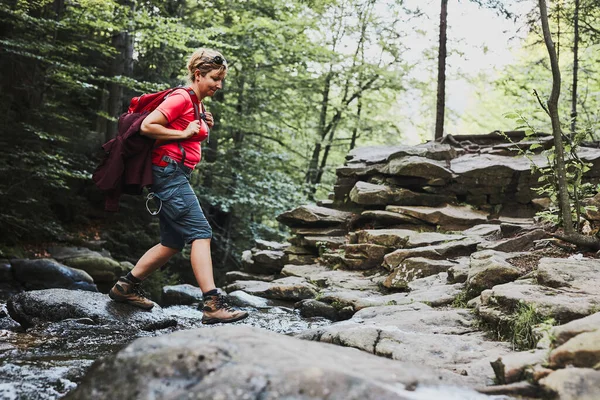 Sırt çantalı bir kadın dağlarda yürüyüş yapıyor, yaz tatilini doğaya yakın geçiriyor. Kadın dereyi geçiyor.