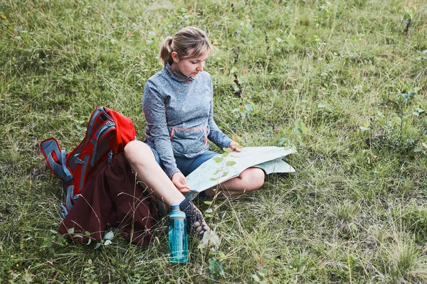 夏休みの日に草の上に座って地図を見て山の中で旅行中に休憩を持っているバックパックを持つ女性 — ストック写真
