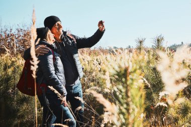 Tatil sırasında selfie çeken bir çift. Sırt çantalı yürüyüşçüler dağlara gidiyor. Güneşli bir günde insanlar çayırda uzun otların arasında yürüyor. Aktif boş zaman doğaya yakın.