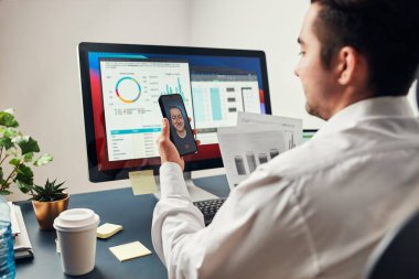 Girişimci adam iş görüşmesi yapıyor, cep telefonuyla görüntülü konuşma yapıyor. Bilgisayar ekranında veri, grafik ve grafiklerle çalışan bir iş adamı. Elinde akıllı telefon tutan adam ofisinde oturuyor.