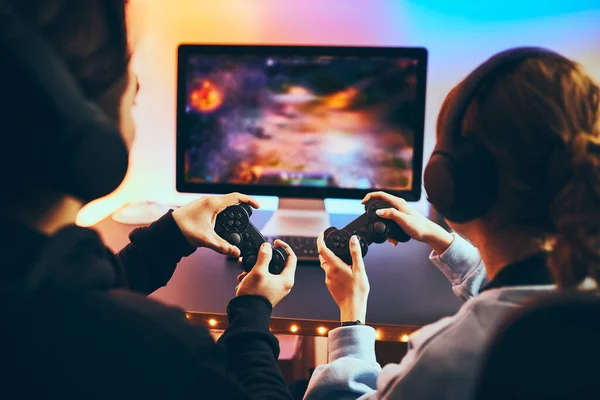 Friends playing video game at home. Gamers holding gamepads sitting at front of screen. Streamers girl and boy playing online in dark room lit by neon lights. Competition and having fun