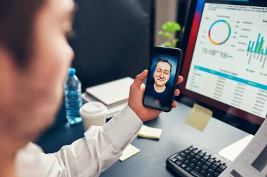 Girişimci adam iş görüşmesi yapıyor, cep telefonuyla görüntülü konuşma yapıyor. Bilgisayar ekranında veri, grafik ve grafiklerle çalışan bir iş adamı. Elinde akıllı telefon tutan adam ofisinde oturuyor.