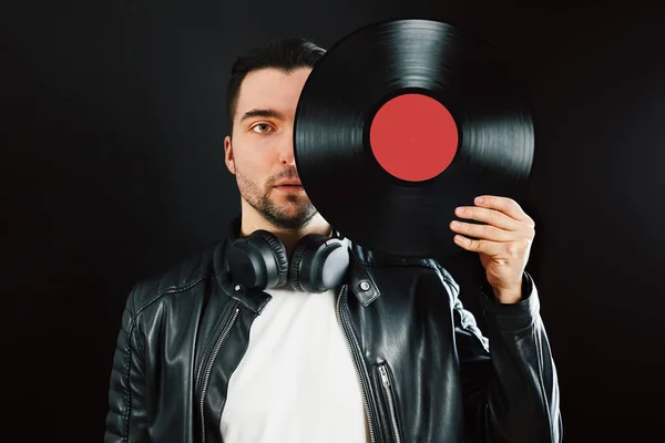 Man holding retro vinyl record covering face. Rock style. Vintage music style. Male wearing black jacket holding vinyl record disc standing on dark background. Retro music. Classic audio