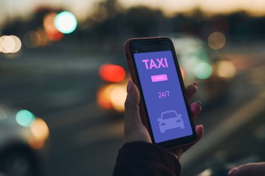 Bir kadın akıllı telefondan mobil uygulama kullanarak internetten taksi çağırıyor. Taksiyi internetten ayırtıyorum. Şehir merkezindeki caddede taksi durağı ayarlıyorum. Araba paylaşımı. Cep telefonunda taksi servisi. Arka planda gece şehir trafik ışıkları
