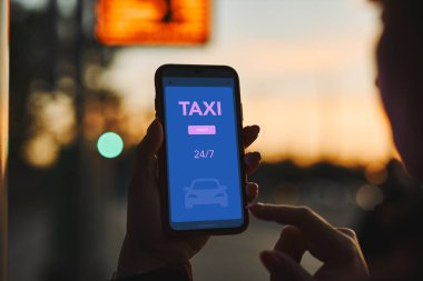 Bir kadın akıllı telefondan mobil uygulama kullanarak internetten taksi çağırıyor. Taksiyi internetten ayırtıyorum. Şehir merkezindeki caddede taksi durağı ayarlıyorum. Araba paylaşımı. Cep telefonunda taksi servisi. Arka planda gece şehir trafik ışıkları
