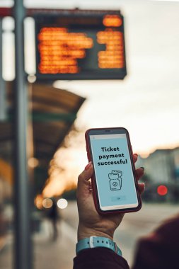 Otobüs durağı yakınlarında bilet alan bir kadın. Bilet parasını mobil ödeme uygulaması ile ödeyen kişi. Hareket halindeki teknolojiyi kullanarak. Seyyar ekrandan onay iste. Uygulamadaki en üst seviye hesap. Arkaplanda otobüs rotaları zamanlaması
