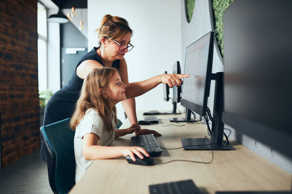 Computer class at school. Teacher assisting schoolgirl while class at primary school. Child learning computer on elementary computer science lesson. Teacher helping to find task solution. Giving clues. Smart little girl learning online at school. Bac