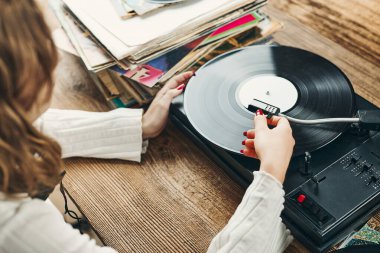 Vinil pikaptan müzik dinleyen genç bir kadın. Pikapta müzik çalıyor. Kadın, evdeki eski plak koleksiyonundan müziğin keyfini çıkarıyor. Analog vinil plak yığını. Retro ve klasik müzik tarzı. Müzik tutkusu ve hobisi