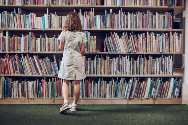 Schoolgirl choosing book in school library. Smart girl selecting literature for reading. Books on shelves in bookstore. Learning from books. School education. Benefits of everyday reading. Child curiosity