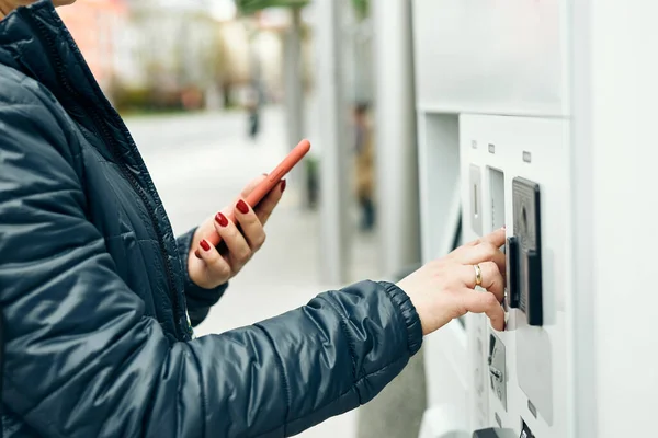 Woman buying ticket at ticket machine paying using mobile payment app on smartphone. Female buying bus public transpoort ticket in vending machine. Person purchasing car parking ticket using mobile phone