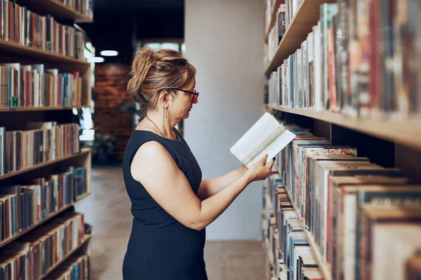 Woman reading book in public library. Teacher searching for literature for reading and learning. Woman standing among bookshelves. Benefits of everyday reading. Books on shelves in bookstore