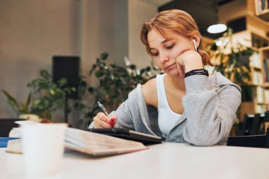 Student learning in university library. Young woman writing essay and making notes using computer tablet. Focused student studying for college exams. Using technology