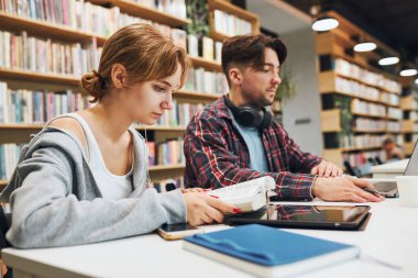 Students learning in university library. Young man preparing for test on laptop. Girl reading textbook. Focused students studying for college exams. Students discussing and learning together sitting at desk. Back to school
