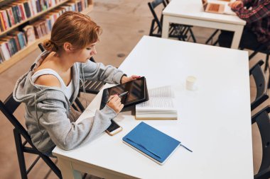 Student learning in university library. Young woman writing essay and making notes using computer tablet. Focused student studying for college exams. Using technology. Back to school