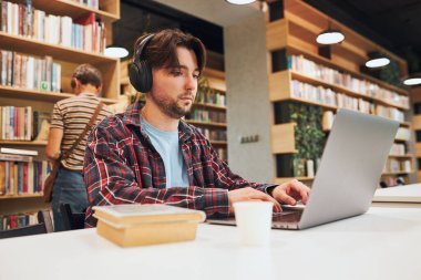 Üniversite kütüphanesinde öğrenci öğrenimi. Genç adam laptopta test için hazırlanıyor. İnternet kursunu dinleyen bir adam. Odaklanmış öğrenci üniversite sınavlarına çalışıyor. Teknoloji kullanılıyor