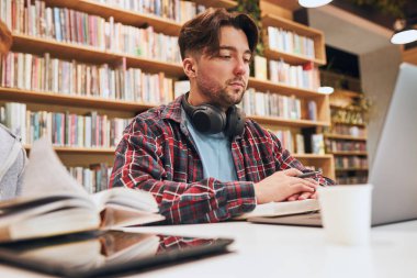 Students learning in university library. Young man preparing for test on laptop. Girl learning from book. Focused students studying for college exams. Students discussing and learning together sitting at desk. Back to school