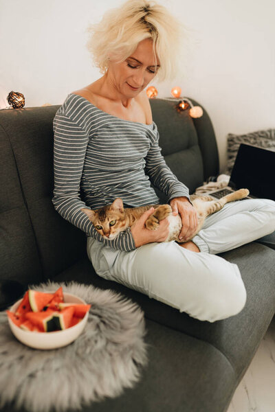 Adult blonde woman sitting on a sofa and playing with a cat