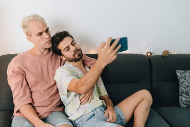 Gay happy couple sitting on sofa and making selfie in their house