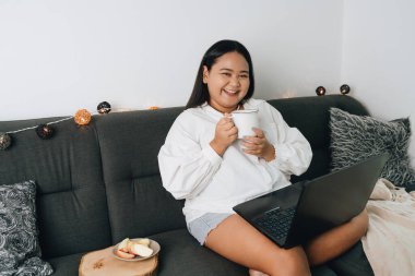 Kanepede oturan ve evde dizüstü bilgisayar kullanan genç bir kadının portresi.