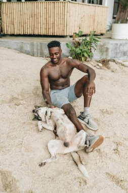 Gülümseyen çekici genç siyah adam sahilde bir köpekle oynuyor. Yüksek kalite fotoğraf
