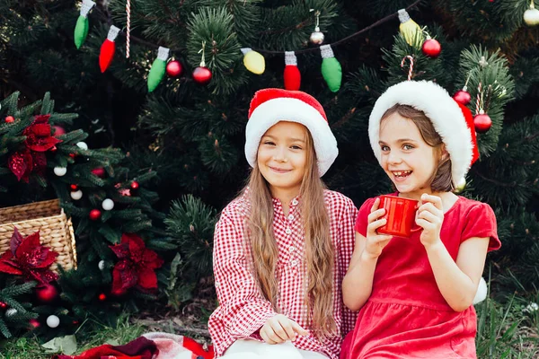 メリークリスマス。ポートレートの二つの面白い子供たち女の子でサンタ帽子食べるジンジャーブレッドクッキー飲むホットチョコレート — ストック写真