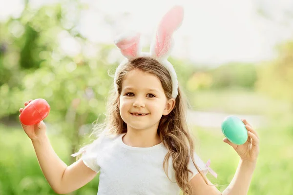 Милая забавная девушка с пасхальными яйцами и кроличьими ушами в саду. Пасхальная концепция. Смеющийся ребенок во время охоты за пасхальными яйцами — стоковое фото