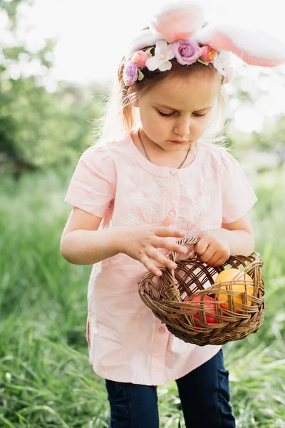 Милая забавная девушка с пасхальными яйцами и кроличьими ушами в саду. Пасхальная концепция. Смеющийся ребенок во время охоты за пасхальными яйцами — стоковое фото