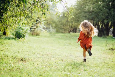 Çocuk döner, çayırda dans eder. Kız doğayla eğleniyor. Gün batımı parkında kaçan sevimli uzun saçlı sarışın kız. Seçici odaklanma. Mutlu bir çocukluk. Boşluğu kopyala