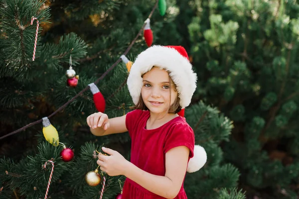 Christmas in july. Child waiting for Christmas in wood in summer. portrait of little girl in red dress decorating christmas tree. winter holidays and people concept. Merry Christmas and Happy Holidays