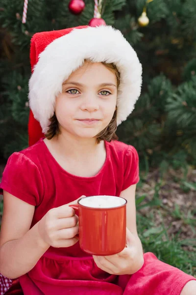 メリークリスマス サンタの帽子の面白い子供の女の子の肖像は 楽しい時間を過ごして外でホットチョコレートを飲むジンジャーブレッドクッキーを食べています ハッピー ホリデー 休日を楽しんでる 7月のクリスマス — ストック写真