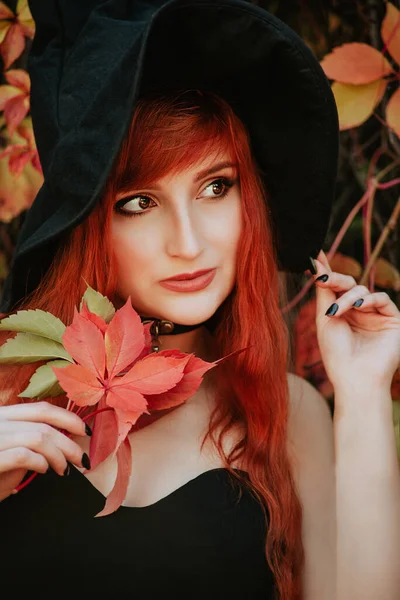 ハロウィーン不気味な暗い魔法の森の背景に魔女の帽子の美しい若い魔女の女の子 秋の明るい葉を持つモデルの女の子の肖像画 秋の外観スタイル 大きな目の魔法の光景を持つ赤毛の女の子 — ストック写真