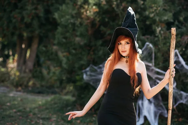 万圣节漂亮的年轻女巫戴着女巫帽戴着恐怖的黑暗魔法森林背景 用雕刻的南瓜 红头发姑娘 大眼睛 迷人的视力 — 图库照片
