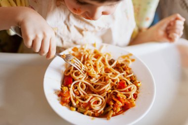 Çocuk sebzeli spagetti yiyor. Çocuk yemek yerken eğleniyor. Yüzü sos kaplı kahverengi saçlı çocuk. Hafta sonu, mutfakta sıcak ve samimi bir sahne.