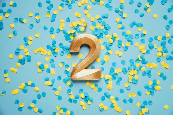 Number 2 two golden celebration birthday candle on yellow and blue confetti Background. two years birthday. concept of celebrating birthday, anniversary, important date, holiday