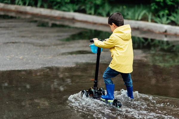 Little boy in raincoat and rubber boots playing in puddle. Fun on street. body hardening in summer. Splashes, drops of water, outdoor. waterproof boots jump in puddle and mud in the rain.
