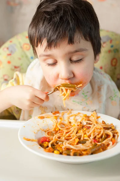 Child eating spaghetti with vegetables. Kid having fun eating. Brown haired boy with face covered in sauce. Weekend, warm and cozy scene in the kitchen