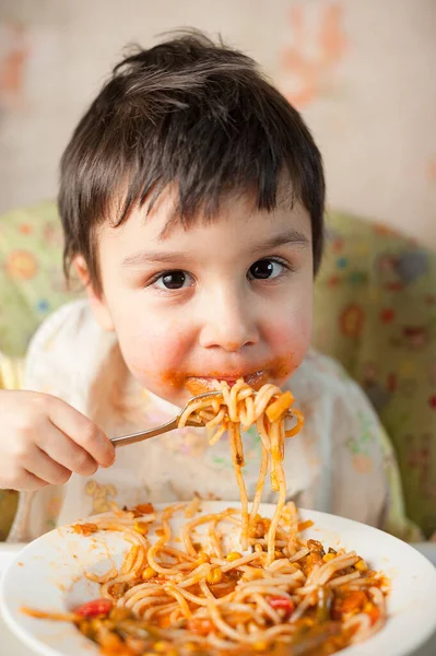 Child eating spaghetti with vegetables. Kid having fun eating. Brown haired boy with face covered in sauce. Weekend, warm and cozy scene in the kitchen