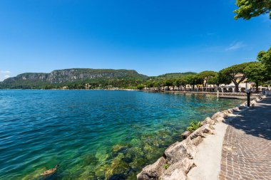 Garda 'da Garda Gölü kıyısındaki göl kıyısı, köy ve turizm beldesi boyunca gezinti alanı (Lago di Garda). Verona, Veneto, İtalya, Avrupa. Ufukta San Vigilio Promontory görünüyor.