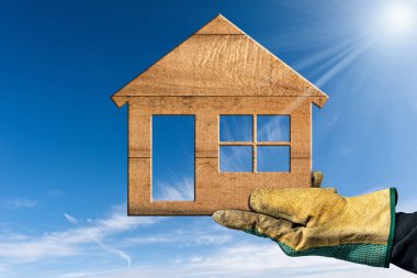 Küçük ahşap bir evi tutan koruyucu iş eldivenli bir elin yakın çekimi bulutlu, güneş ışınları ve kopyalama alanı olan açık mavi bir gökyüzüne karşı. Eko ev kavramı.