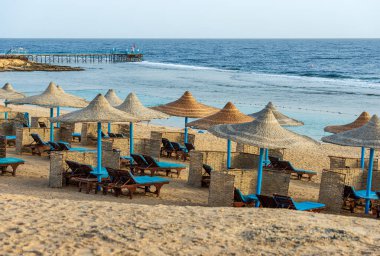 Marsa Alam, Mısır, Afrika yakınlarında, Kızıl Deniz 'in kumlu plajı, saman şemsiyeli ve şezlonglu. Mercan resifinde deniz manzarası ve dalgalar kırılıyor..